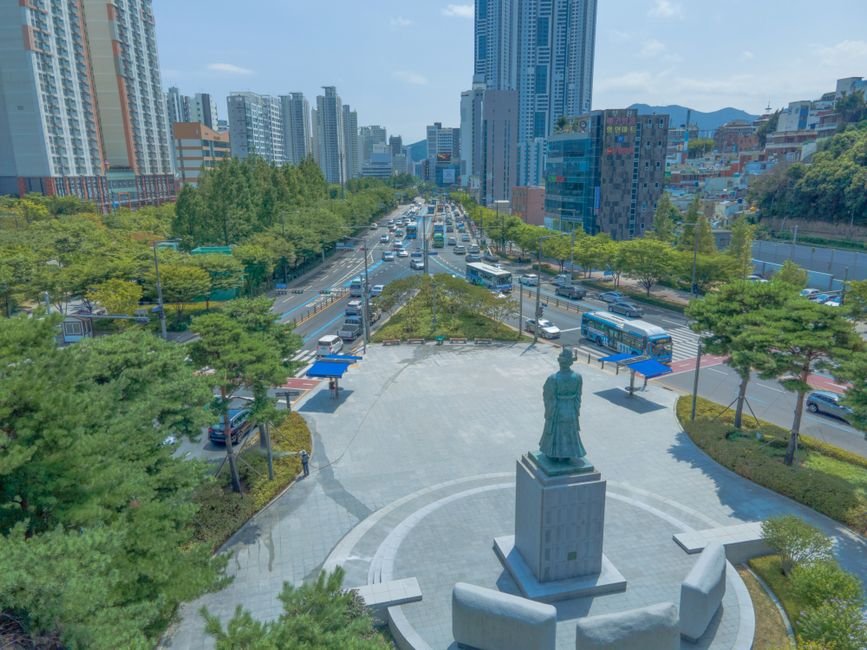 부산 도심의 분주한 항공 뷰로 도시 경관과 조각상을 보여줍니다.